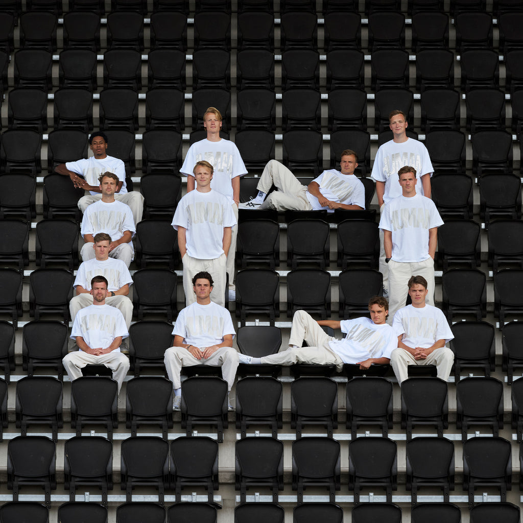 Multiple men from the Dutch national hockey team in white 10D FOR HUMAN shirts and beige pants sitting on black stadium chairs.