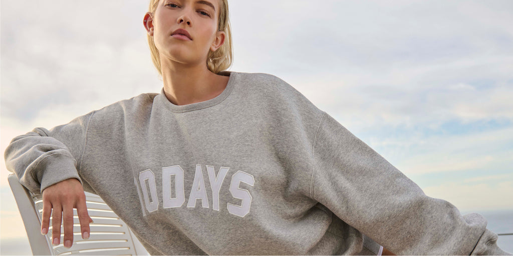 Woman wearing the light grey melee 10DAYS statement sweater, sitting on a white chair near the sea.