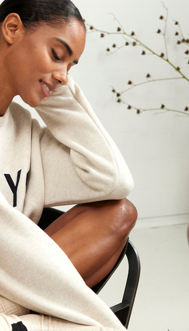 Woman sitting in chair, wearing a soft white melee statement sweater. 