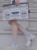 Person carrying a silver radio, walking in a light background.